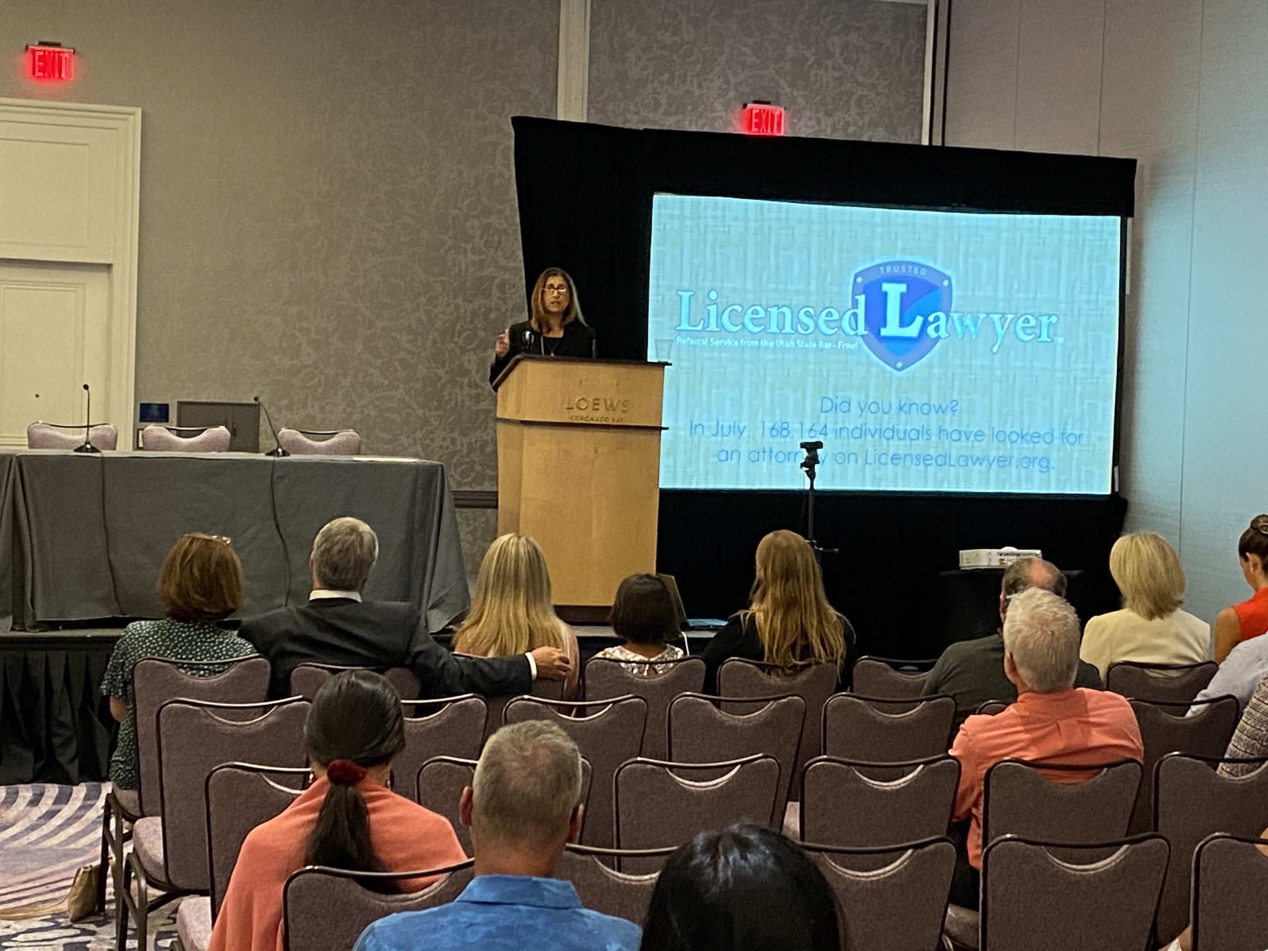 Female at podium presenting to a seated audience with a screen in the background showing the home page of LicensedLawyer.org